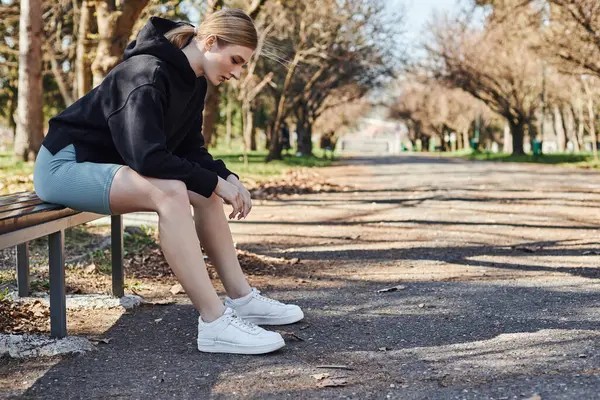 Loira e apto jovem mulher em preto com capuz desportivo e shorts de ciclismo sentado no banco de madeira no parque — Fotografia de Stock