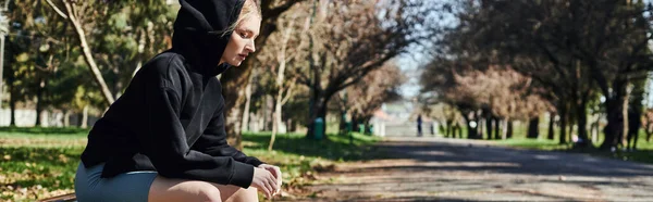 Fit jeune femme en sweat à capuche sport noir et short cycliste assis sur un banc en bois dans le parc, bannière — Photo de stock