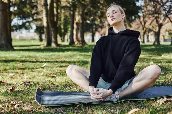 Forme jeune femme aux cheveux blonds et vêtements de sport assis sur tapis de fitness et méditer dans le parc — Photo de stock