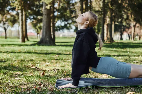 Подходит молодая женщина с светлыми волосами и спортивной одеждой медитирует и тянется назад на коврик для йоги в парке — стоковое фото