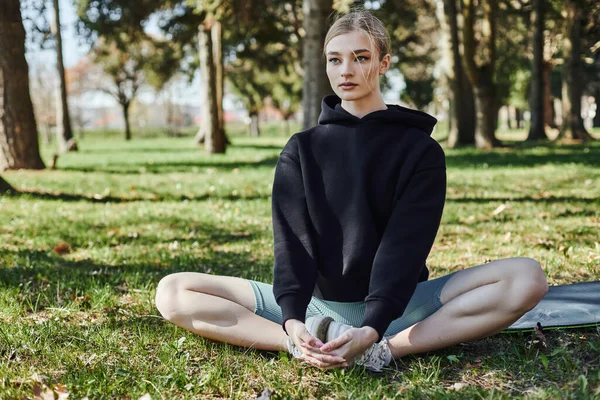 Спортивная молодая женщина со светлыми волосами и спортивной одеждой сидит на коврике и медитирует в парке — стоковое фото