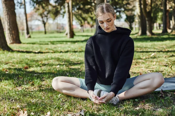 Fit young woman with blonde hair and sportswear sitting on sports mat and meditating in park — Stock Photo