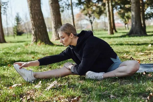 Mujer joven en forma con el pelo rubio y ropa deportiva estiramiento mientras está sentado en la alfombra de fitness en el parque - foto de stock