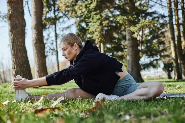 Fit young woman with blonde hair and sportswear stretching leg while sitting on fitness mat in park — Stock Photo