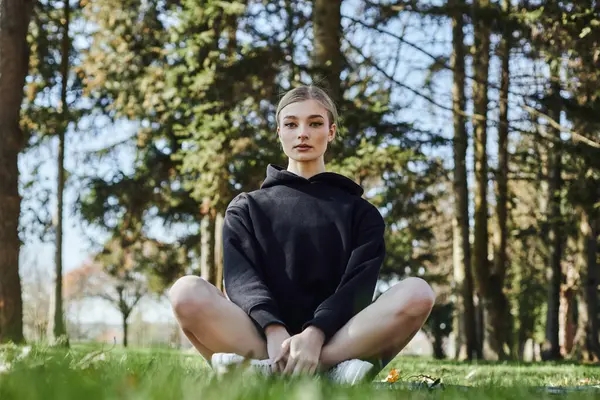 Красивая и стройная женщина с светлыми волосами и спортивной одеждой, сидящая на коврике во время медитации в парке — стоковое фото