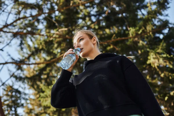 Mujer joven en forma con el pelo rubio y ropa deportiva beber agua dulce después de hacer ejercicio en el parque - foto de stock