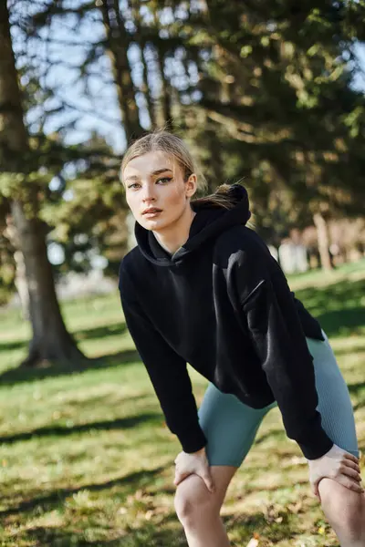 Forme jeune femme avec des cheveux blonds et des vêtements de sport travaillant tout en regardant la caméra dans le parc — Photo de stock
