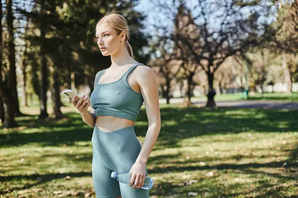 Sportive blonde en tenue active tenant bouteille d'eau et smartphone dans le parc, la motivation et le sport — Photo de stock