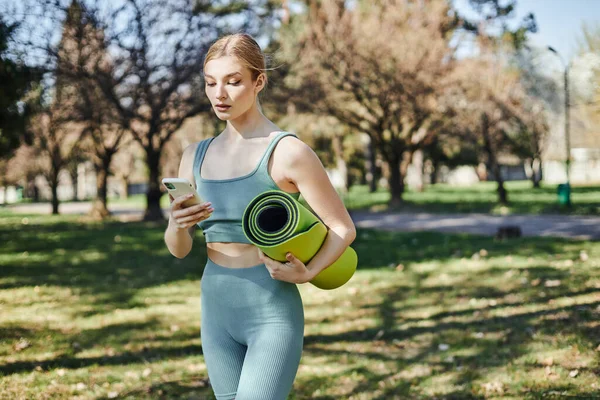 Bonita esportista em desgaste ativo segurando smartphone e tapete de fitness enquanto está de pé no parque — Fotografia de Stock