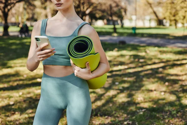 Ausgeschnittene Ansicht einer Sportlerin in aktiver Kleidung mit Smartphone und Fitnessmatte im grünen Park — Stockfoto