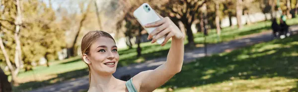Счастливая спортсменка в активной одежде делает селфи на смартфоне после тренировки в парке, баннер — стоковое фото