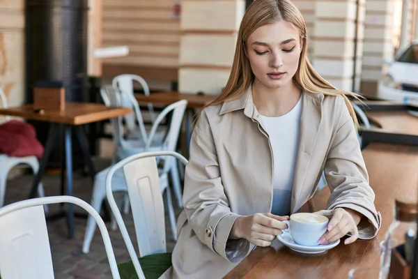Blonde junge Frau im schicken beigen Trenchcoat mit Blick auf ihren Cappuccino im Café, Tasse Kaffee — Stockfoto