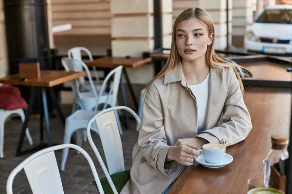 Красивая молодая женщина в стильном бежевом плаще смотрит на капучино в кафе, чашку кофе — стоковое фото