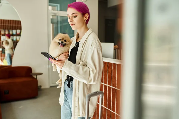 Femme avec message spitz poméranienne sur smartphone près de la réception et valise à l'hôtel pour chien — Photo de stock