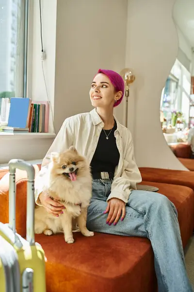 Smiley woman looking away while sitting with pomeranian spitz near suitcase in cozy pet hotel — Stock Photo