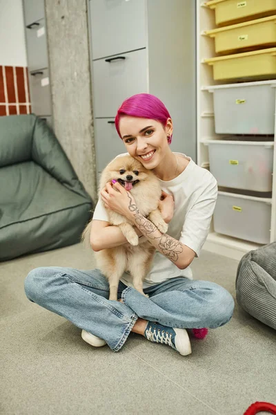 Niñera de mascotas tatuada con pelo morado abrazando al perro esponjoso y sonriendo a la cámara en el piso en el hotel de mascotas - foto de stock
