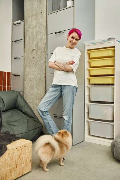 Cuidador tatuado y de pelo morado con los brazos cruzados sonriendo a la cámara cerca de lindo perrito en hotel de mascotas — Stock Photo