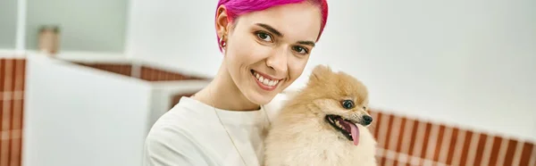 Joyful purple-haired dog sitter with cute pomeranian spitz looking at camera in pet hotel, banner — Stock Photo