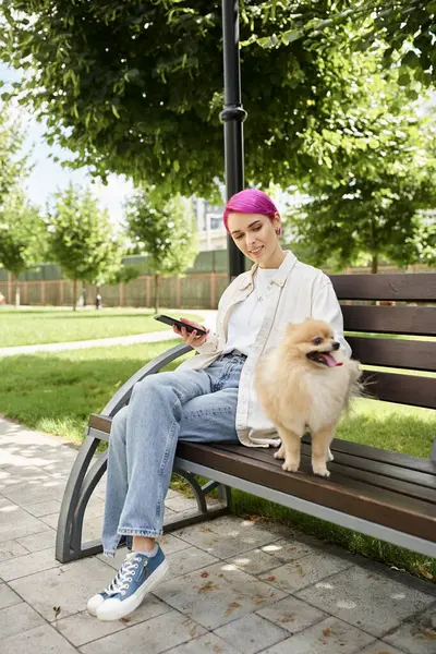 Улыбающаяся пурпурная женщина со смартфоном, сидящая на скамейке в парке рядом со смешным померанским шпионом — стоковое фото