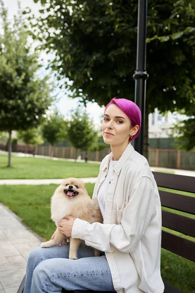 Веселая фиолетовая женщина с померанским шпицем сидит на скамейке в парке и смотрит в камеру — стоковое фото