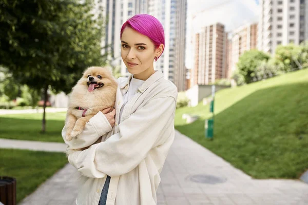 Cheerful purple-haired woman holding funny pomeranian spitz in hands and looking at camera on street — Stock Photo