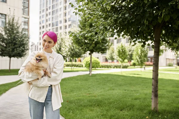 Happy woman with trendy hairstyle walking on city street with pomeranian spitz in hands, enjoyment — Stock Photo