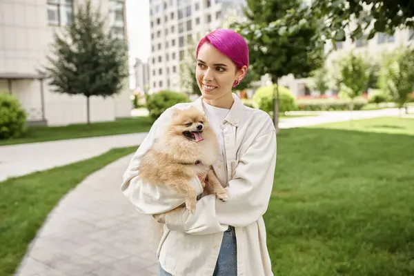 Glückliche lila-haarige Frau hält flauschigen Hund in den Händen und schaut weg auf Straße, Stadtspaziergang — Stockfoto