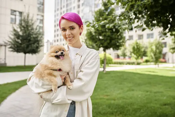 Delighted purple-haired woman with pomeranian spitz in hands looking at camera on street, city life — Stock Photo