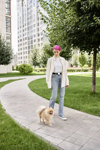 Полная длинноволосая женщина с фиолетовыми волосами, идущая с геранским шпицем по городской улице, весело улыбается. — стоковое фото