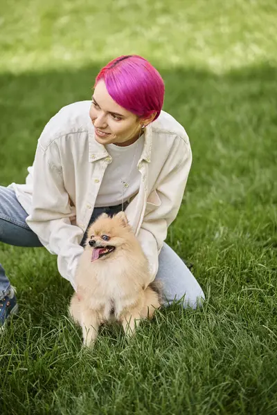 Allegra donna dai capelli viola coccole divertente pomerania spitz e guardando lontano su erba verde nel parco — Foto stock