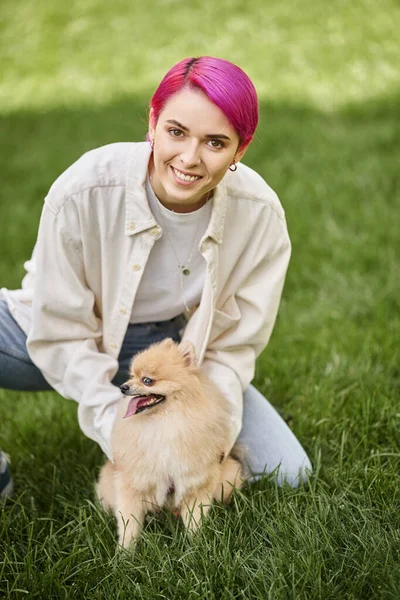 Joyful purple-haired woman embracing funny pomeranian spitz on grassy lawn and smiling at camera — Stock Photo
