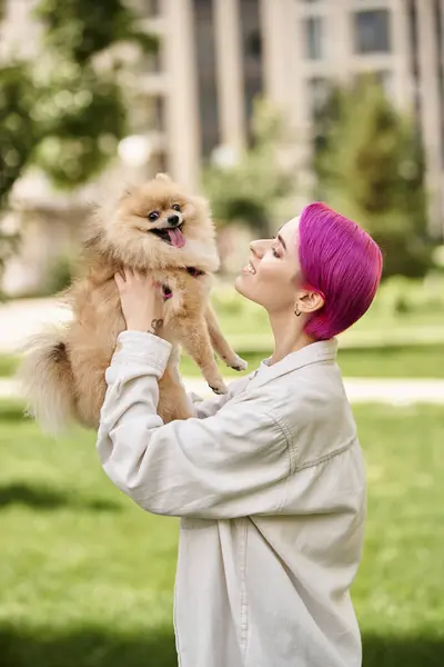 Seitenansicht einer fröhlichen Frau mit lila Haaren, die im Park entzückende Pommernspitzen in den Händen hält — Stockfoto