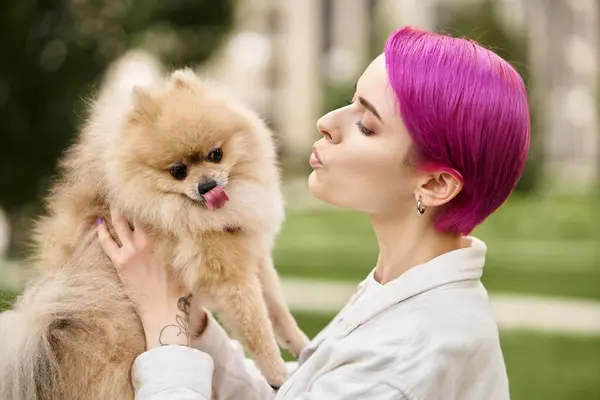 Trendy woman with purple hair pouting lips while holding adorable pomeranian spitz outdoors — Stock Photo