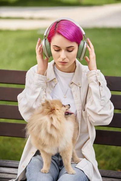 Purple haired woman listening music in wireless headphones while sitting with dog on park bench — Stock Photo