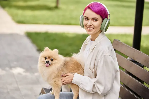 Unbekümmerte lila-haarige Frau mit Kopfhörern sitzt mit pelziger Freundin auf Bank und lächelt in die Kamera — Stockfoto