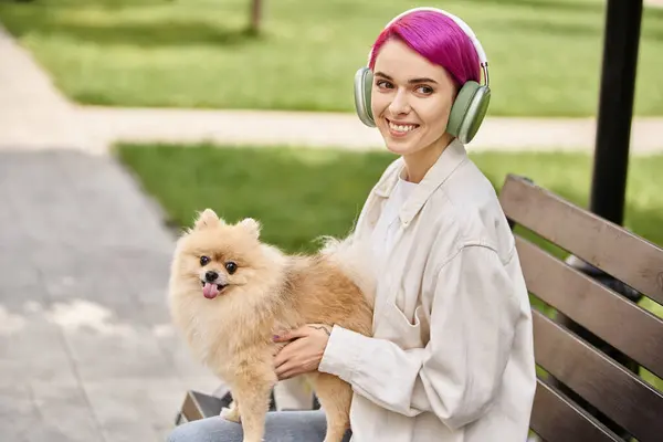 Spensierata donna dai capelli viola in cuffia seduta sulla panchina con amico peloso e sorridente alla macchina fotografica — Foto stock