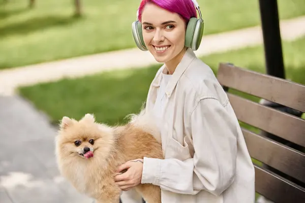 Довольная пурпурная женщина, сидящая в парке с померанским шпицем и слушающая музыку в наушниках — стоковое фото
