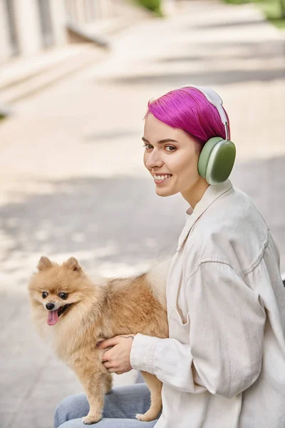 Glückliche lila-haarige Frau mit Kopfhörern, die wegschaut, während sie mit einem flauschigen Freund im Park sitzt — Stockfoto