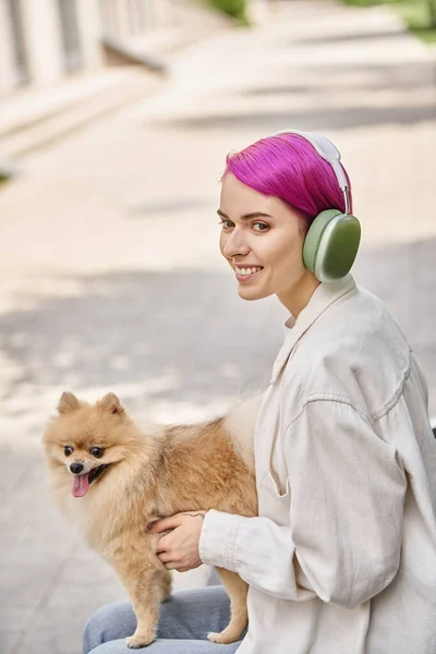 Happy purple-haired woman in headphones looking away while sitting with fluffy friend in park — Stock Photo