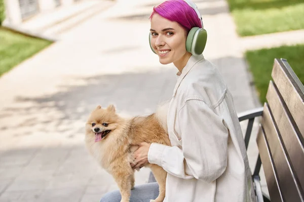 Frau mit lila Haaren sitzt mit Hund im Park und hört Musik über Kopfhörer — Stockfoto