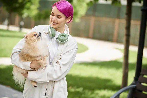 Positive purple-haired woman with headphones holding pampered pomeranian spitz in hands outdoors — Stock Photo