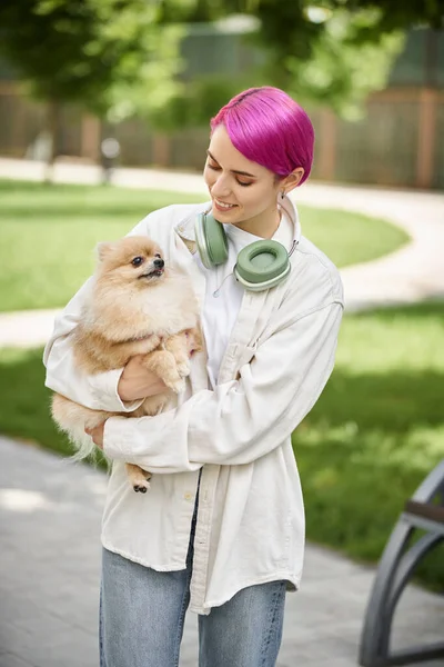 Pleased woman with purple hair and headphones standing with loveable pomeranian spitz in hands — Stock Photo