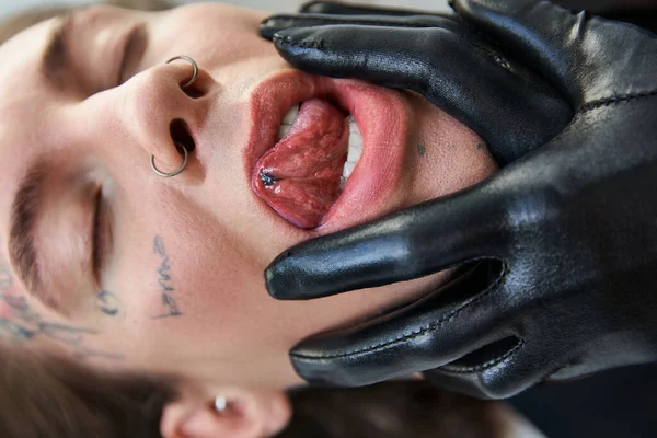 Close up of young man with his tongue out with piercing and tattoos, hand in leather gloves on face — Stock Photo