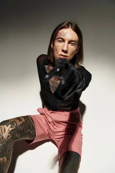 Junger Mann mit Tätowierungen sitzt auf dem Boden und zeigt mit der Hand in Lederhandschuh in die Kamera, Modekonzept — Stockfoto