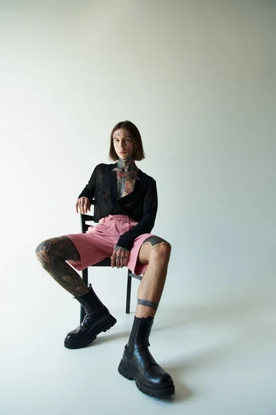 Junger gutaussehender Mann mit Tätowierungen in rosa Shorts sitzt auf Stuhl und schaut in die Kamera, Modekonzept — Stockfoto