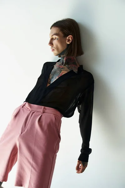 Uomo alla moda in pantaloncini rosa e camicia nera trasparente appoggiata al muro distogliendo lo sguardo, moda — Foto stock