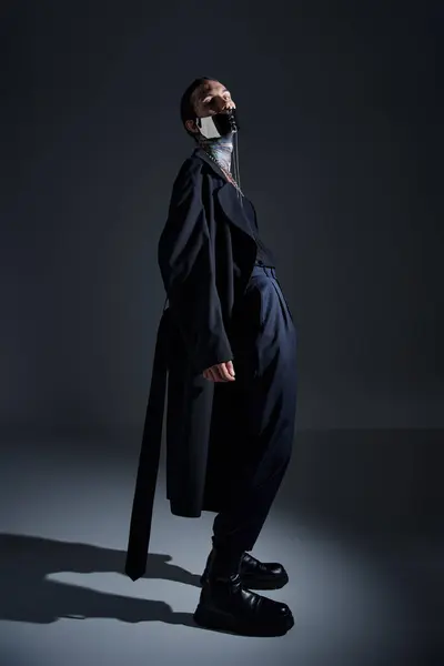 Guapo hombre con estilo en negro abrigo largo y futurista con cordones máscara posando y mirando a la cámara - foto de stock