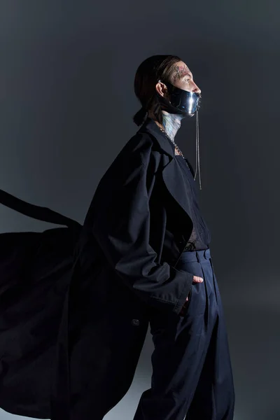 Modelo masculino guapo en abrigo negro y máscara atada futurista posando en perfil con manos en bolsillos - foto de stock
