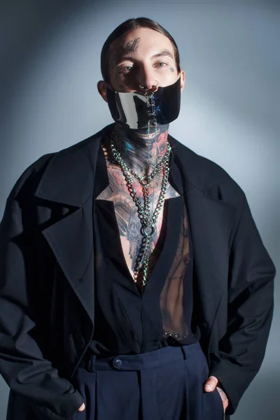 Vertical shot of alluring young man with tattoos and laced mask with accessories looking at camera — Stock Photo