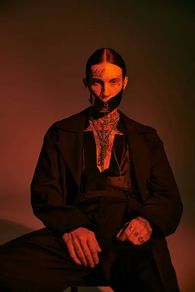 Bonito modelo masculino elegante em traje preto com máscara atada sentado e olhando para a câmera, moda — Fotografia de Stock